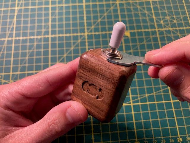 A close-up of a hand holding Focus Switch, a small wooden switch box with a white toggle switch, while another hand uses a small metal tool to adjust or remove the switch. The background is a green cutting mat with yellow grid lines.